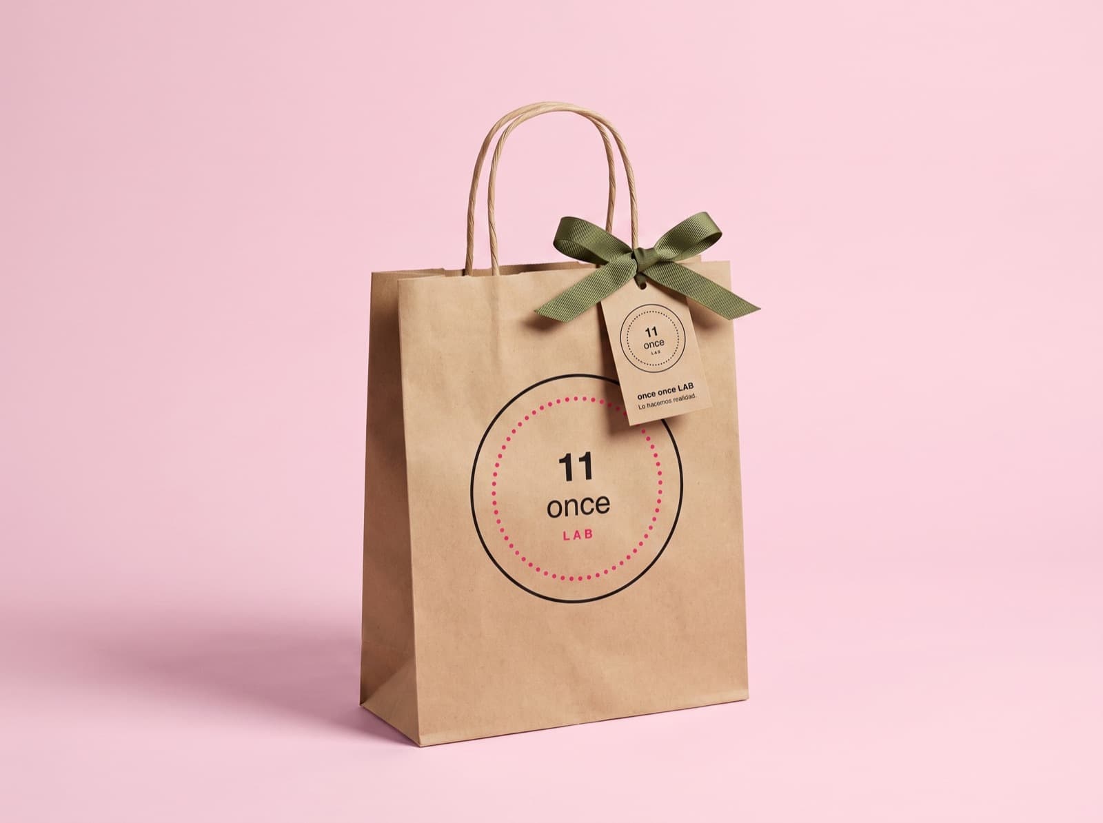 Bolsa de papel kraft con sello circular, moño verde y etiqueta, sobre fondo rosa.