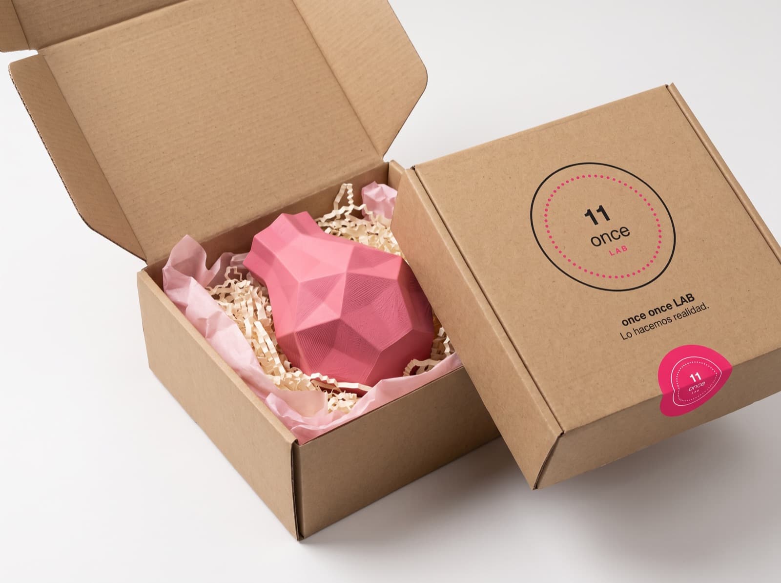 Caja de cartón abierta con una pieza facetada rosa sobre viruta y papel de seda.