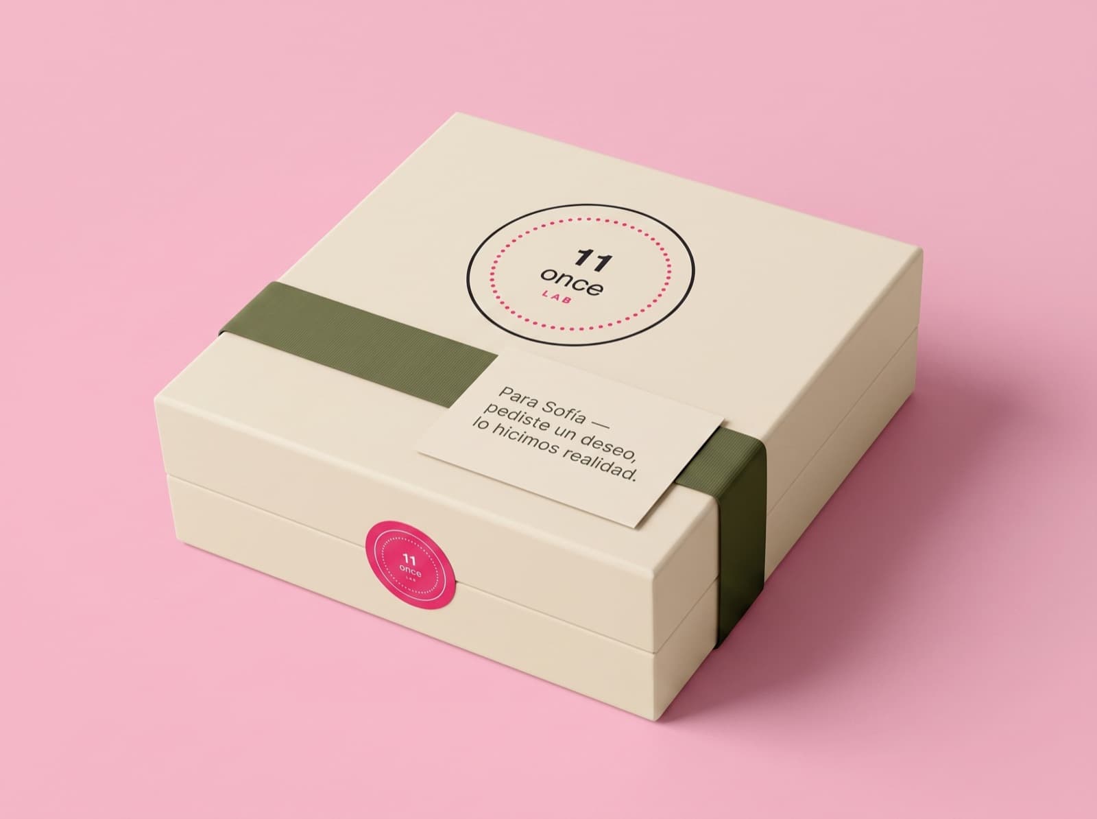 Caja crema con faja verde olivo, tarjeta y sello rosa sobre fondo rosa.