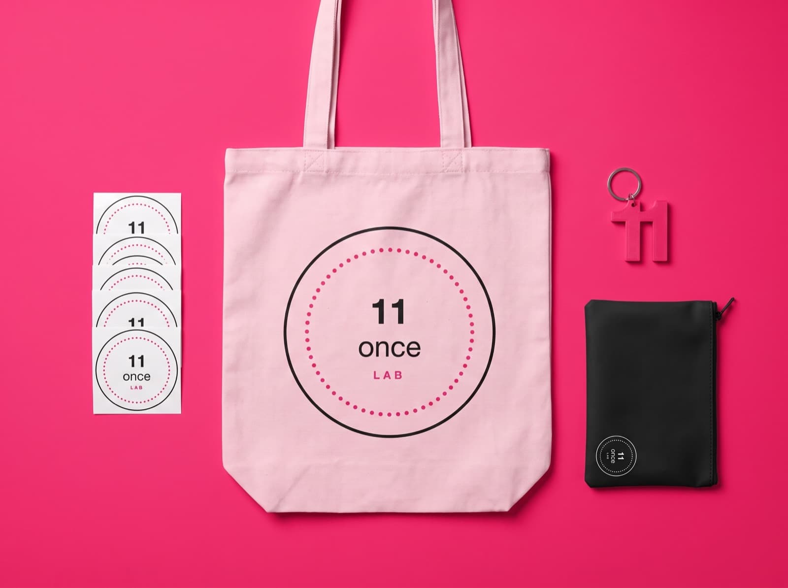 Bolsa de tela rosa con sello circular, llavero, estuche negro y calcomanías sobre fondo rosa.