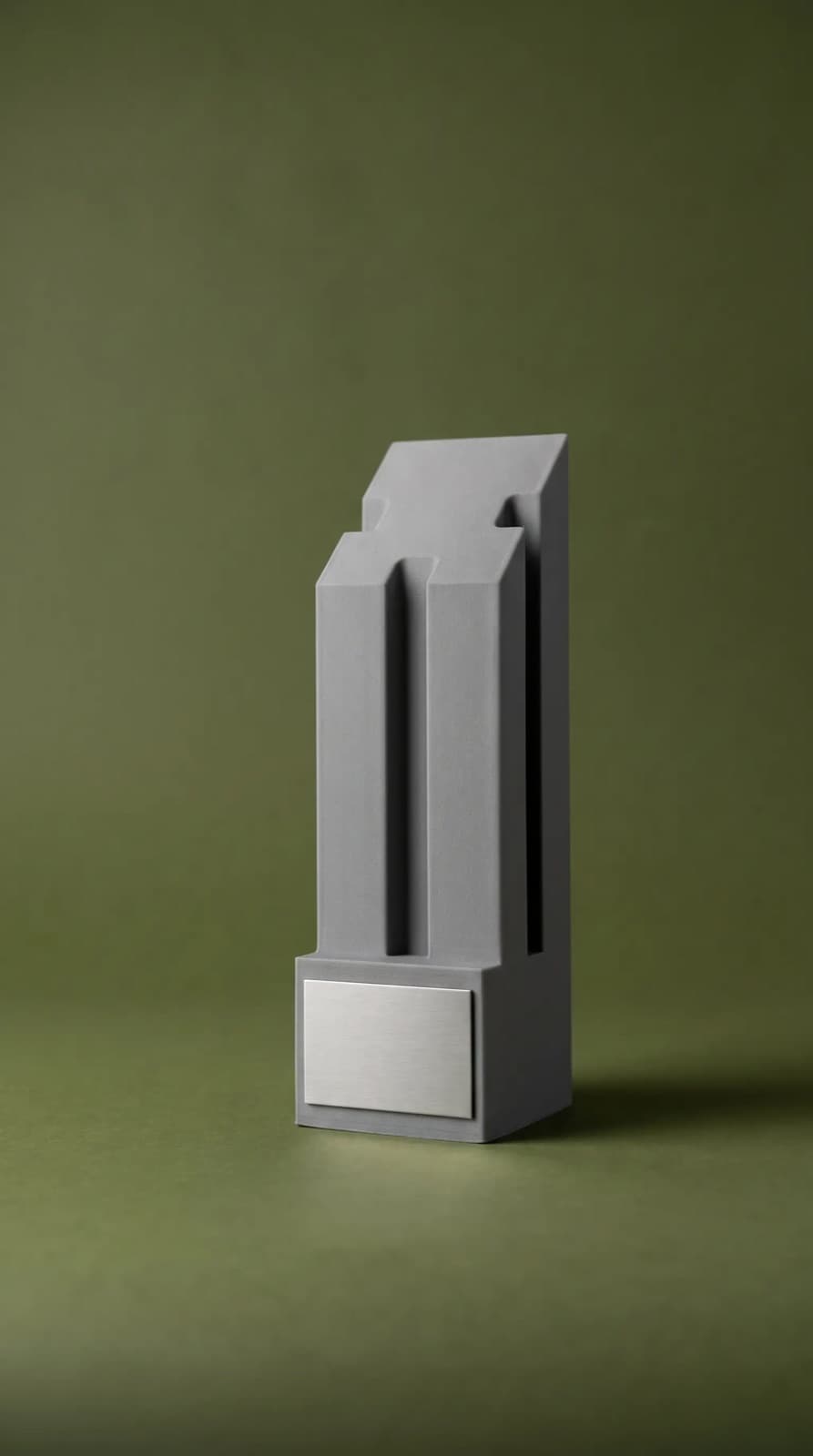 Trofeo gris en forma de torre escalonada con placa plateada sobre fondo verde olivo.